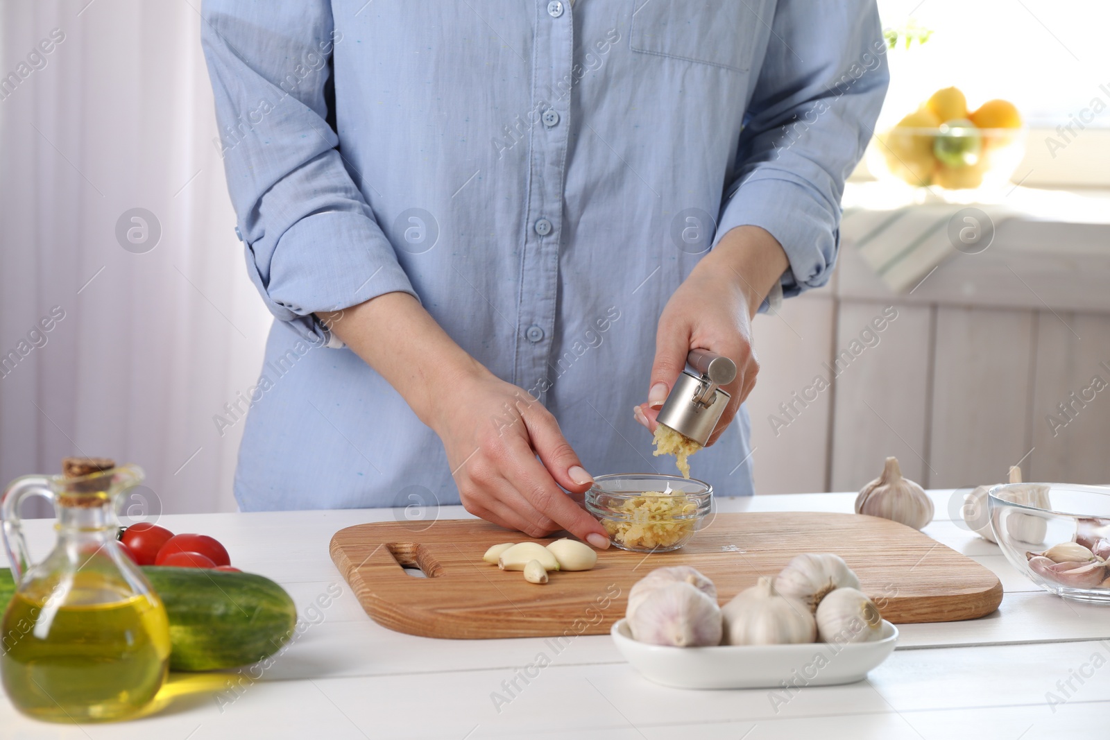 Photo of Woman squeezing garlic with press at white wooden table in kitchen, closeup