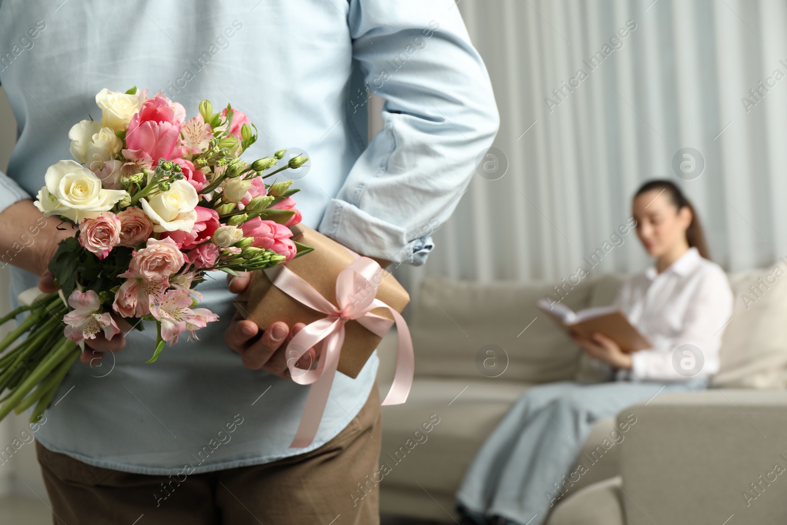 Photo of Man hiding bouquet of flowers and present for his beloved woman indoors, closeup