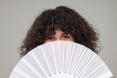 Woman hiding her face behind hand fan on light grey background