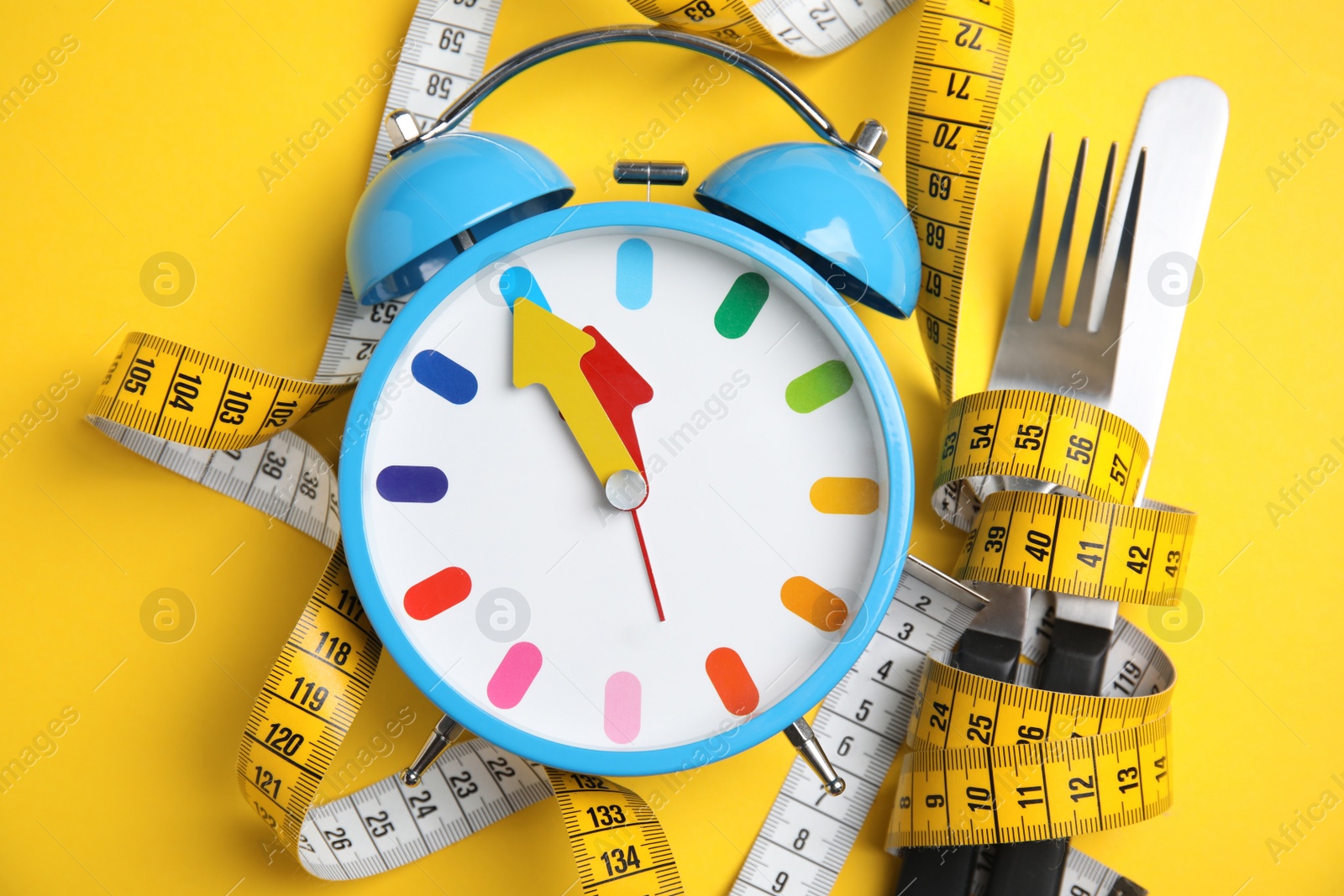 Photo of Alarm clock, measuring tape and cutlery on yellow background, flat lay. Diet regime