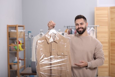 Photo of Dry-cleaning service. Happy man holding hanger with jacket in plastic bag indoors, space for text