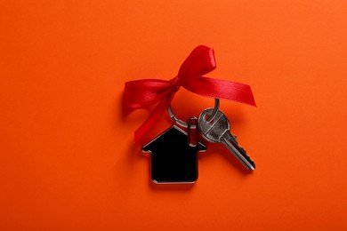 Key with trinket in shape of house and red bow on orange background, top view. Housewarming party