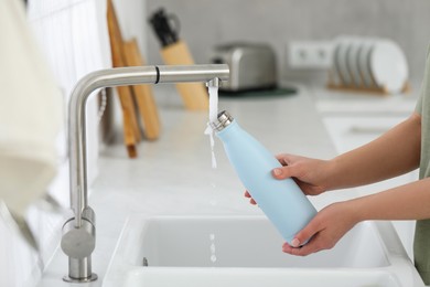 Photo of Woman pouring fresh water from tap into thermo bottle in kitchen, closeup