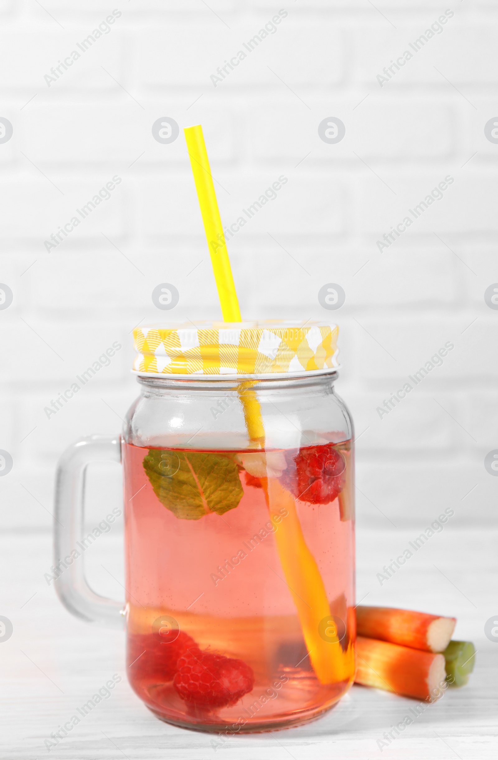 Photo of Mason jar of tasty rhubarb cocktail with raspberry and stalks on white wooden table, closeup