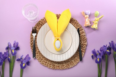 Photo of Festive table setting with painted egg, plates and iris flowers on lilac background, flat lay. Easter celebration