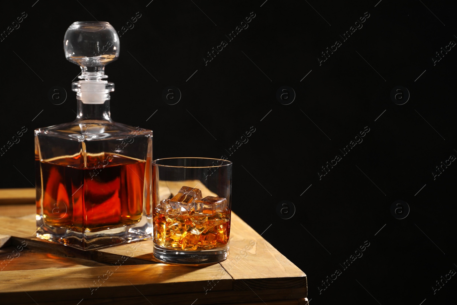 Photo of Whiskey with ice cubes in glass and bottle on wooden crate against black background, space for text