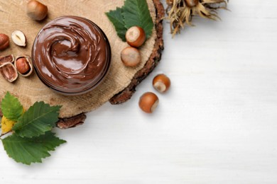 Photo of Jar with tasty chocolate spread and hazelnuts on white wooden table, flat lay. Space for text