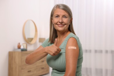 Photo of Senior woman with adhesive bandage on her arm after vaccination showing thumb up at home