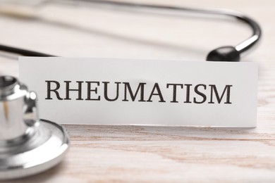 Card with word Rheumatism and stethoscope on white wooden table, closeup