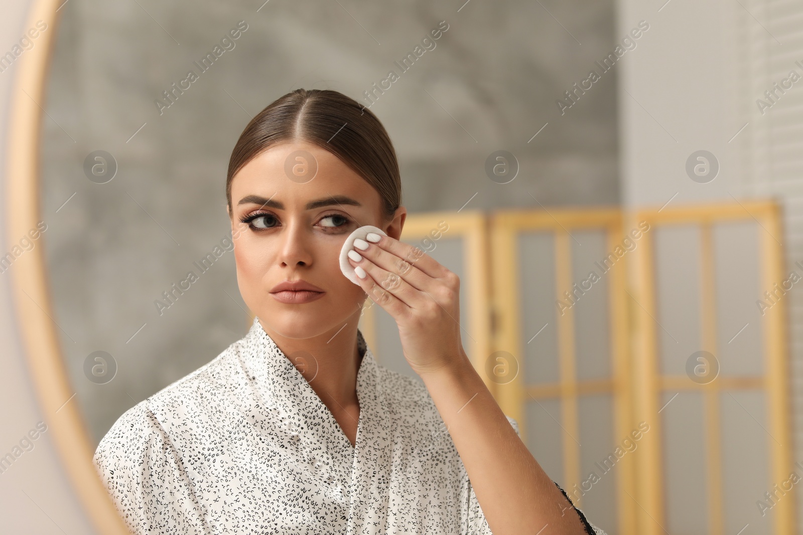 Photo of Beautiful woman removing makeup with cotton pad near mirror indoors, space for text