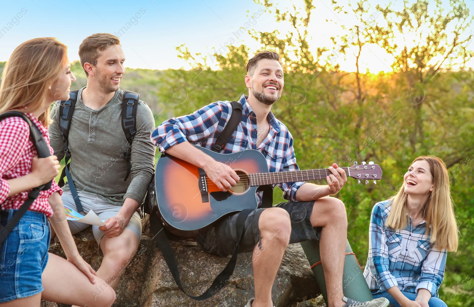 Photo of Young man with backpack playing guitar for his friends in wilderness. Camping season