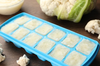 Cauliflower puree in ice cube tray and fresh cauliflower on wooden table, closeup