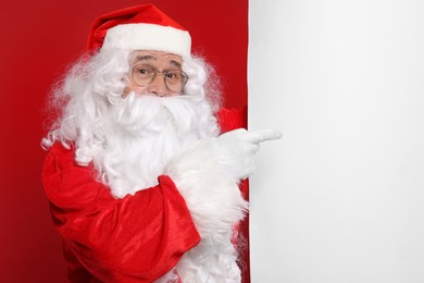 Photo of Santa Claus pointing at blank poster on red background, space for text
