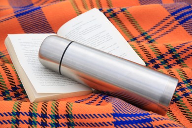 Photo of Metallic thermos with hot drink and open book on plaid, closeup