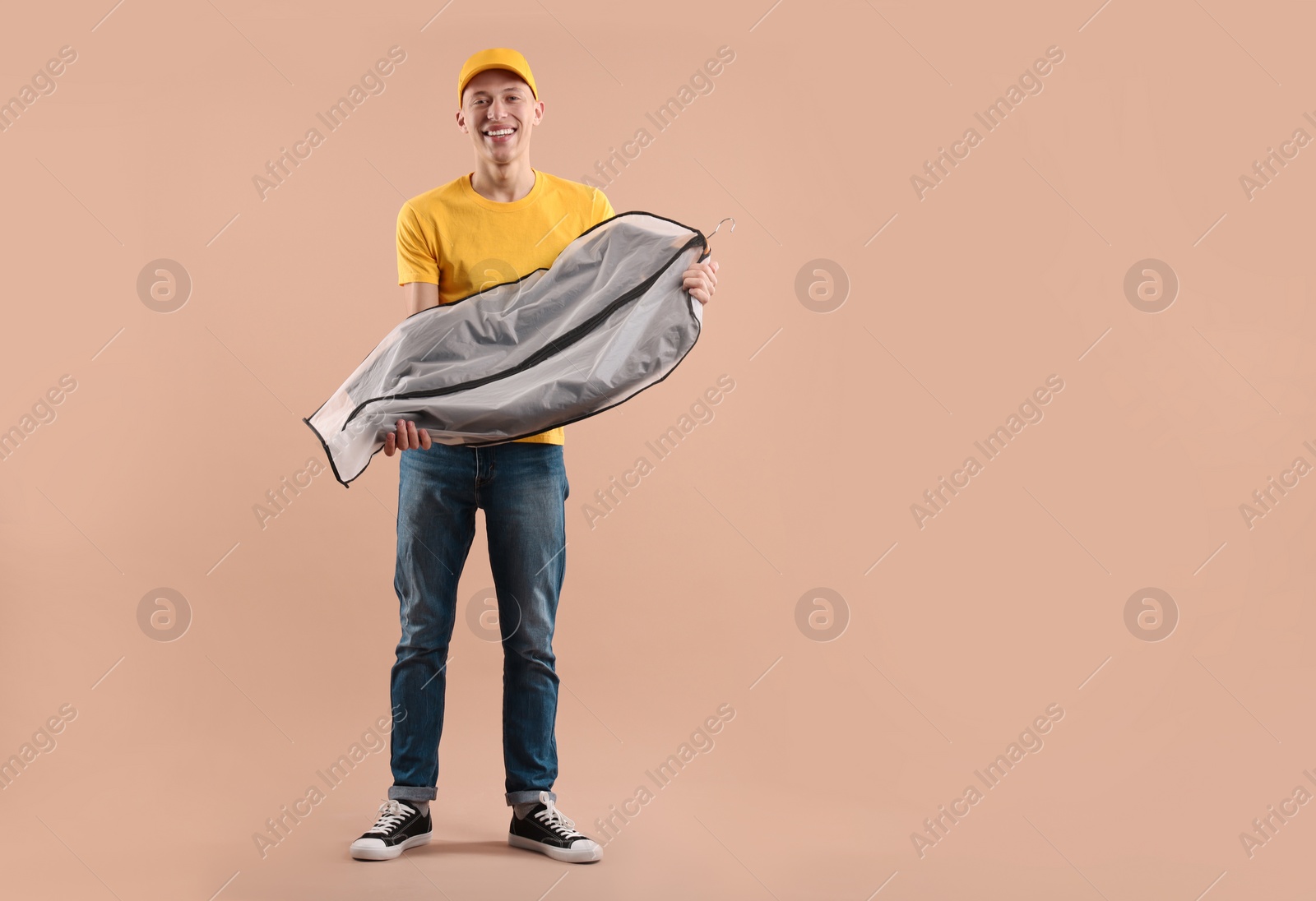 Photo of Dry-cleaning delivery. Happy courier holding garment cover with clothes on beige background, space for text