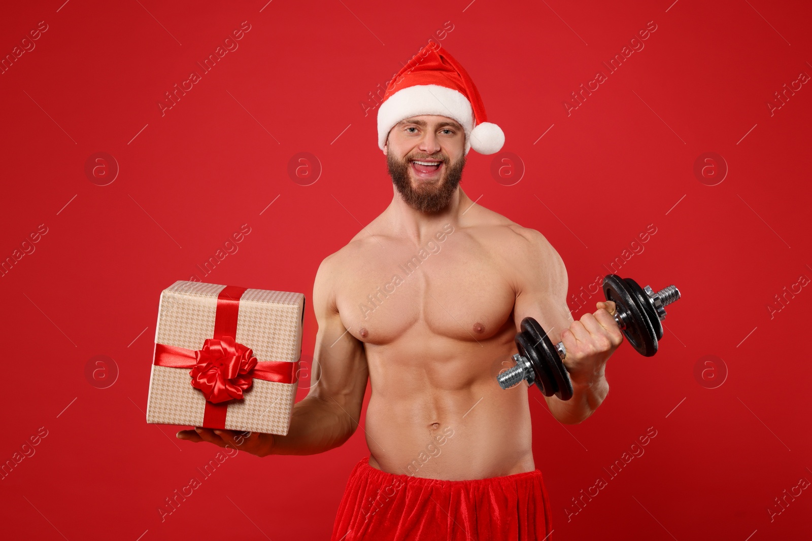 Photo of Muscular young man in Santa hat with dumbbell and Christmas gift box on red background