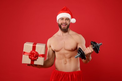 Muscular young man in Santa hat with dumbbell and Christmas gift box on red background