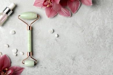 Natural jade face roller, cosmetic product and flowers on grey background, flat lay. Space for text