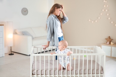 Photo of Cute baby girl in crib and young mother suffering from postnatal depression at home