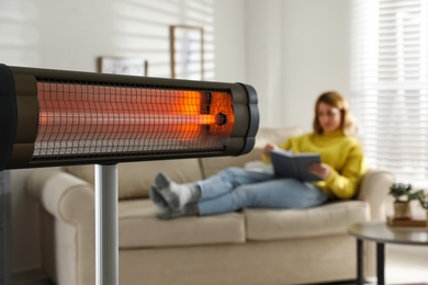 Photo of Woman reading book in living room, focus on electric infrared heater
