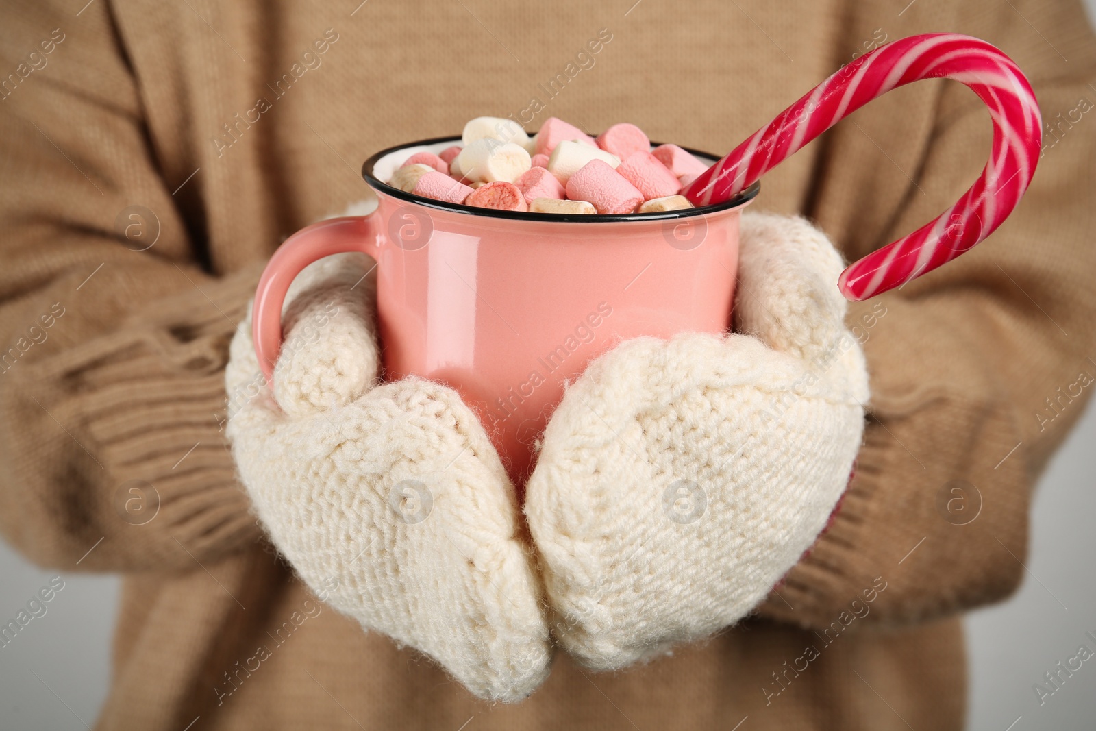 Photo of Woman in knitted mittens holding cup of delicious hot chocolate with marshmallows and candy cane, closeup