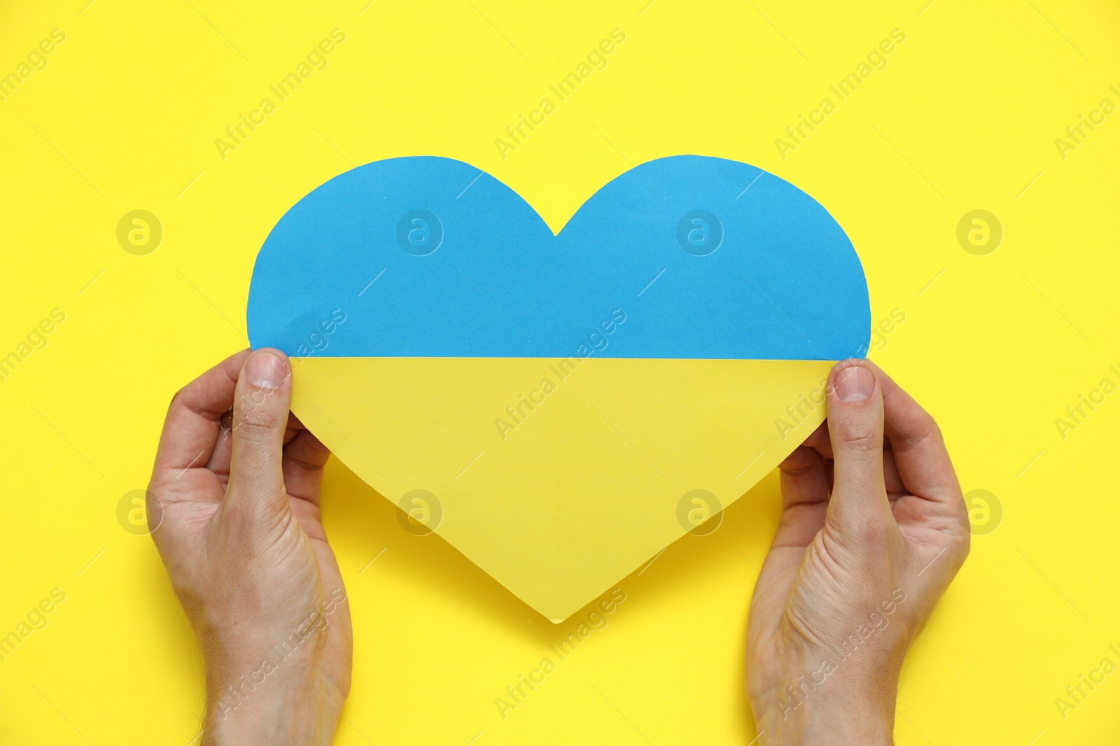 Photo of Man holding paper heart in colors of Ukrainian flag on yellow background, top view