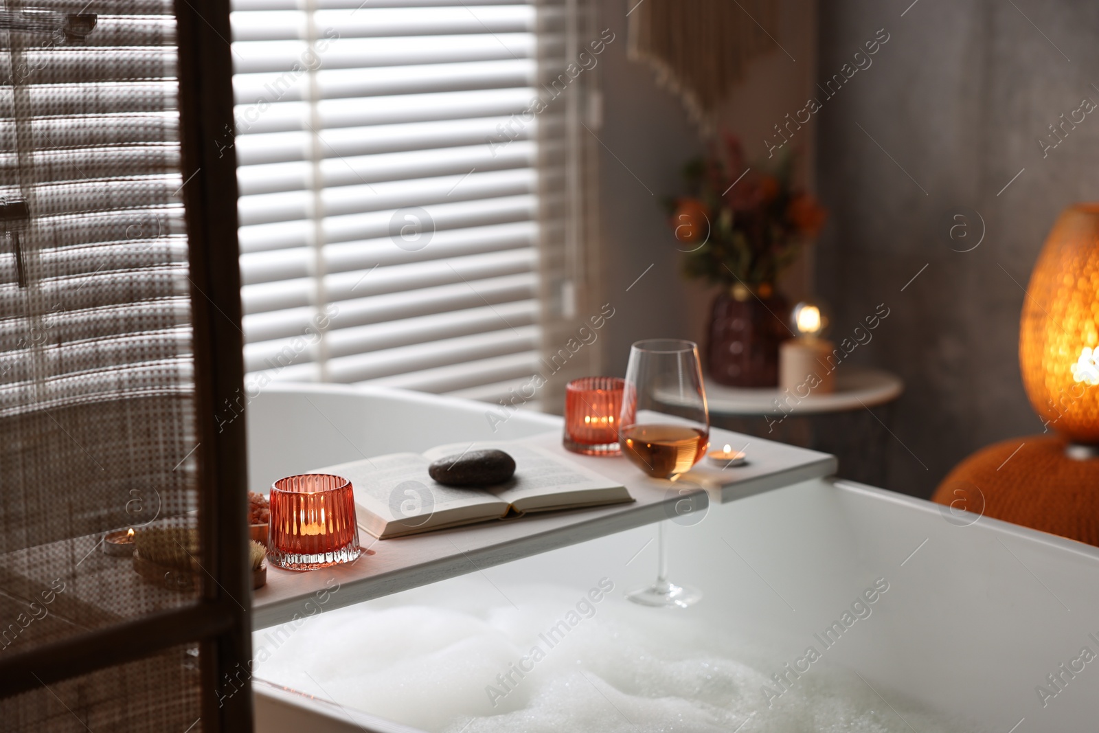 Photo of White wooden tray with glass of rose wine, book and burning candles on bathtub in bathroom