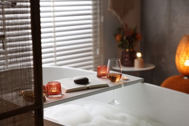 Photo of White wooden tray with glass of rose wine, book and burning candles on bathtub in bathroom