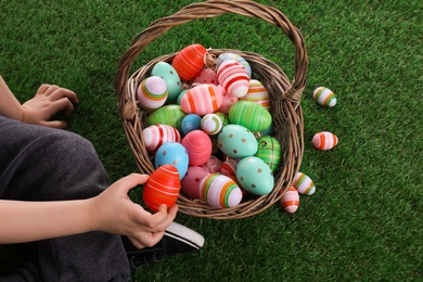 Photo of Little boy with basket full of dyed Easter eggs on green grass, closeup