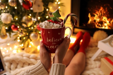 Photo of Woman holding cup of sweet drink near fireplace at home, closeup. Cozy winter holidays atmosphere