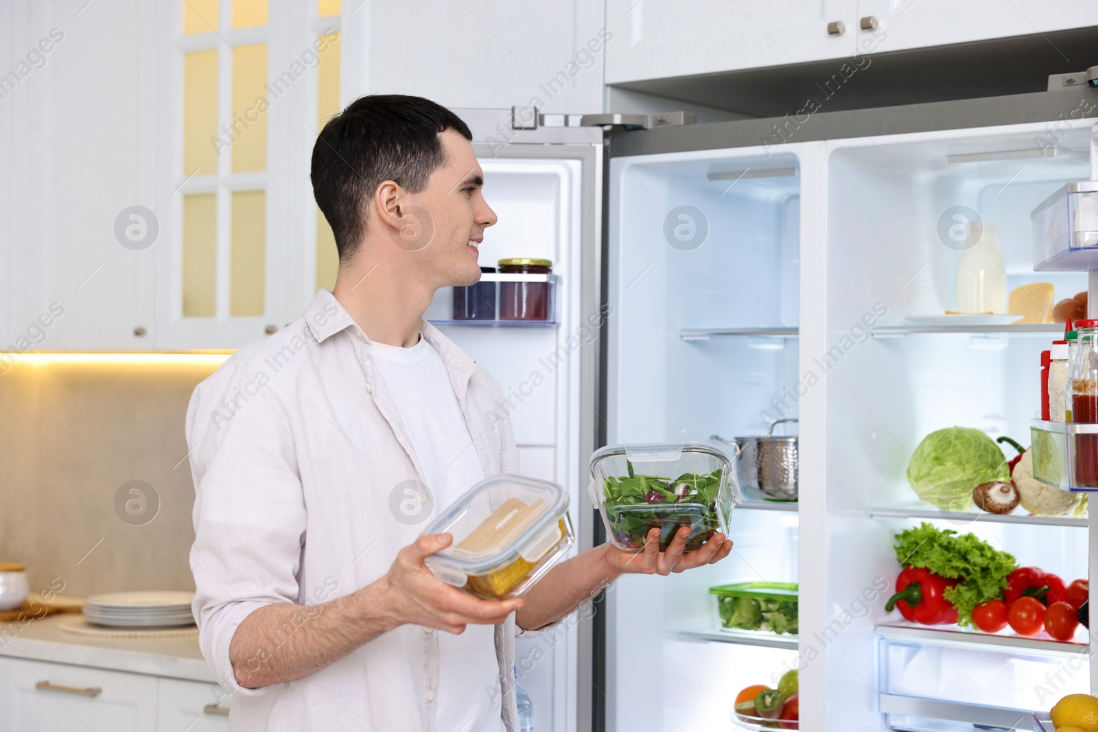 Photo of Happy man holding containers with vegetables near refrigerator in kitchen