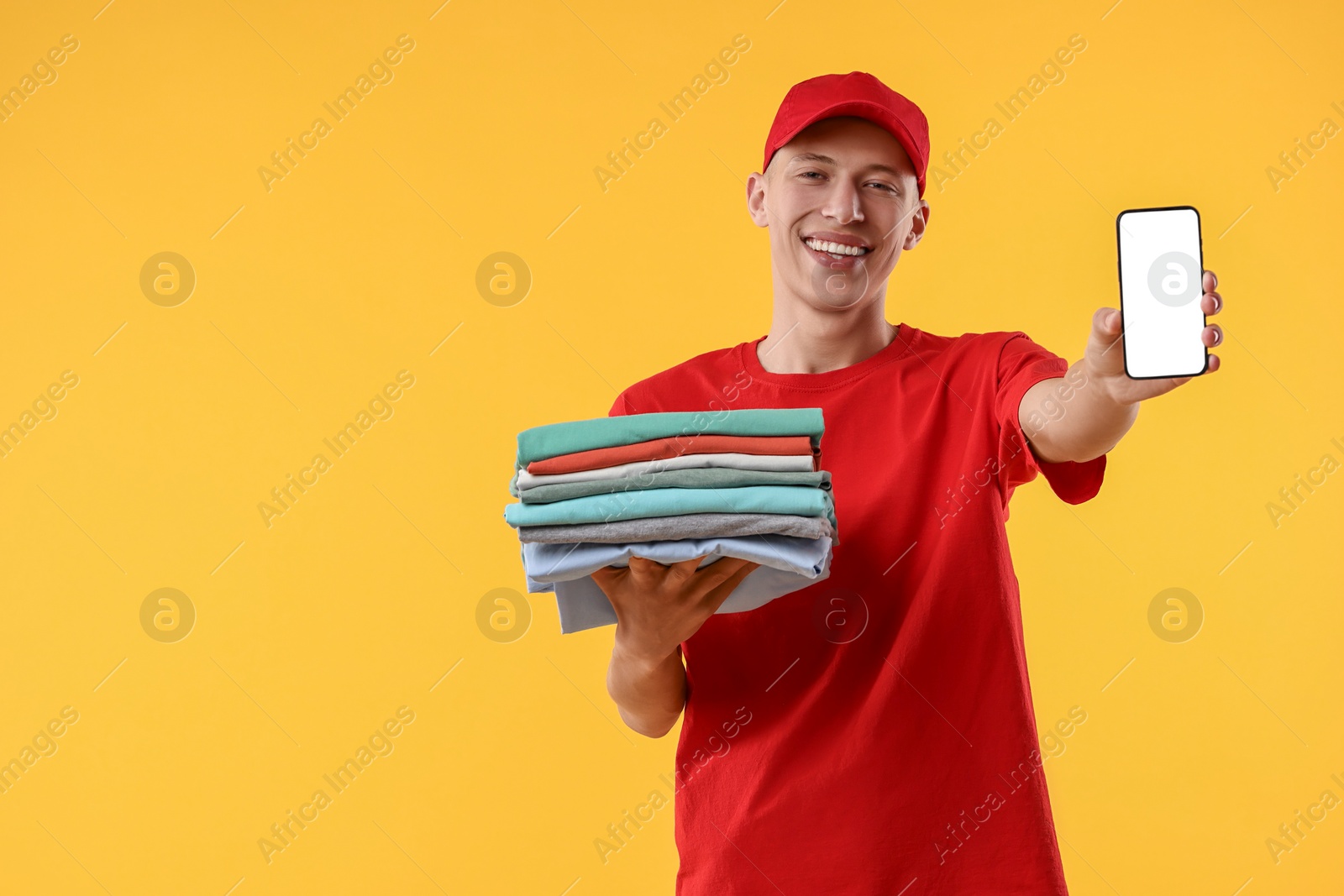 Photo of Dry-cleaning delivery. Happy courier holding folded clothes and smartphone on orange background, space for text