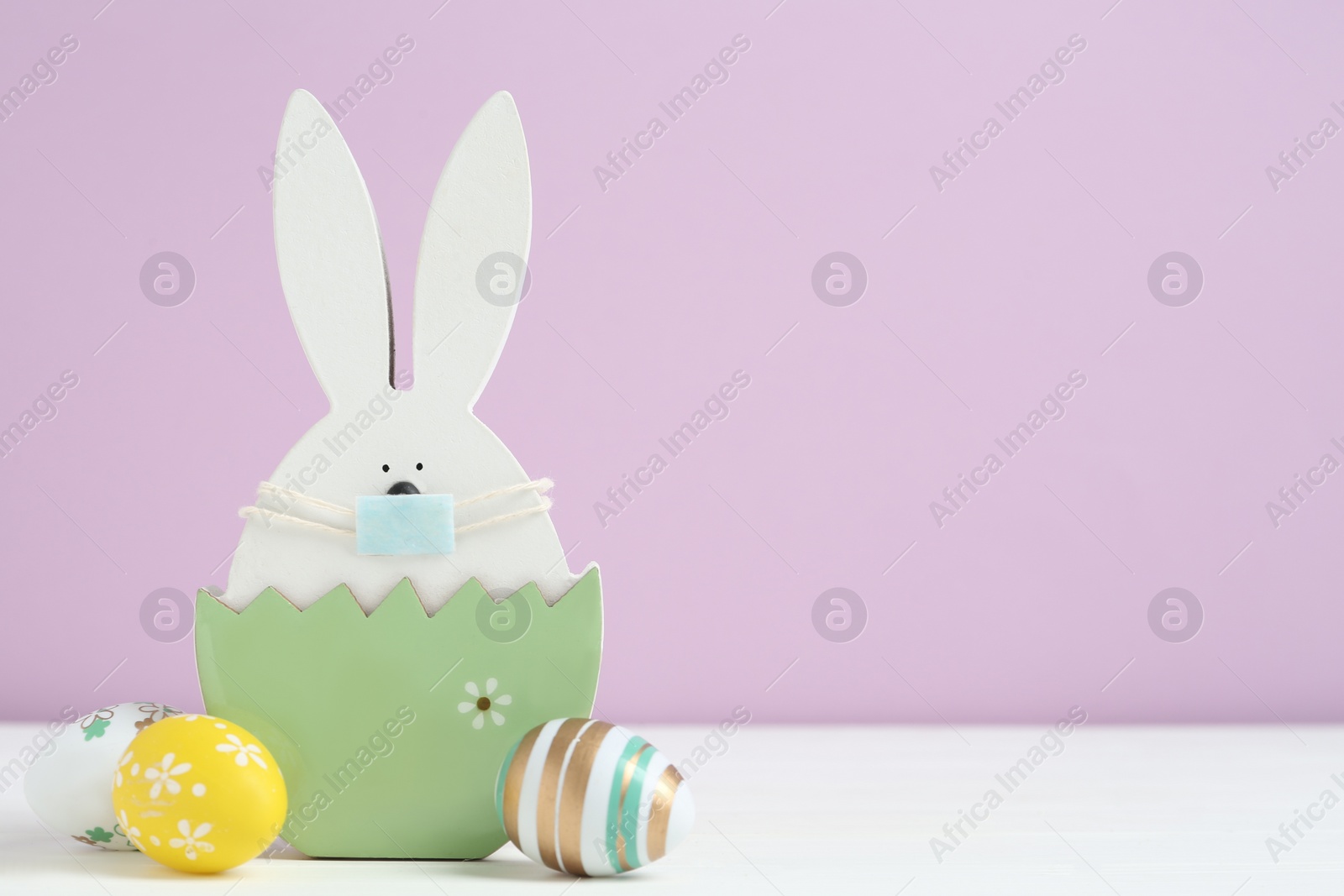 Photo of Wooden bunny with protective mask, painted eggs and space for text on white wooden table. Easter holiday during COVID-19 quarantine