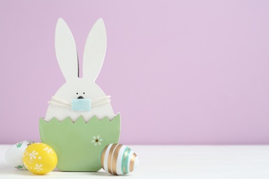 Wooden bunny with protective mask, painted eggs and space for text on white wooden table. Easter holiday during COVID-19 quarantine