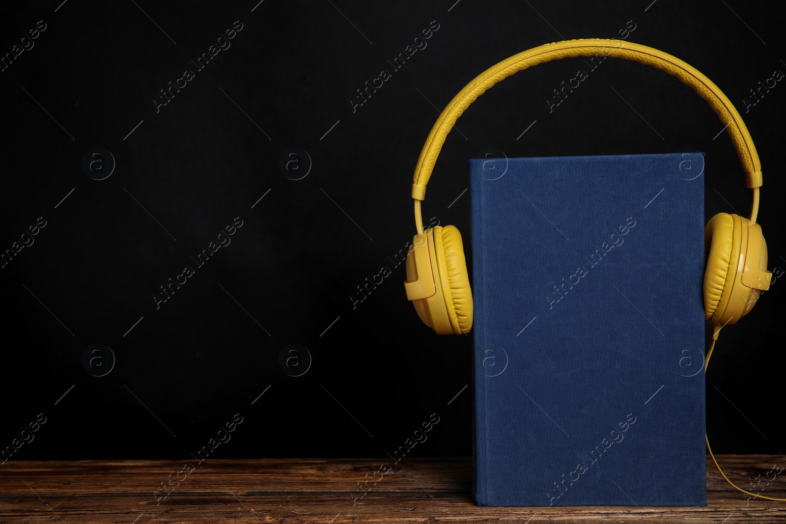 Photo of Book and modern headphones on wooden table against black background. Space for text