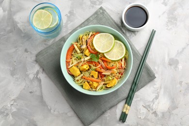 Photo of Stir-fry. Delicious cooked noodles with chicken and vegetables in bowl served on gray textured table, flat lay