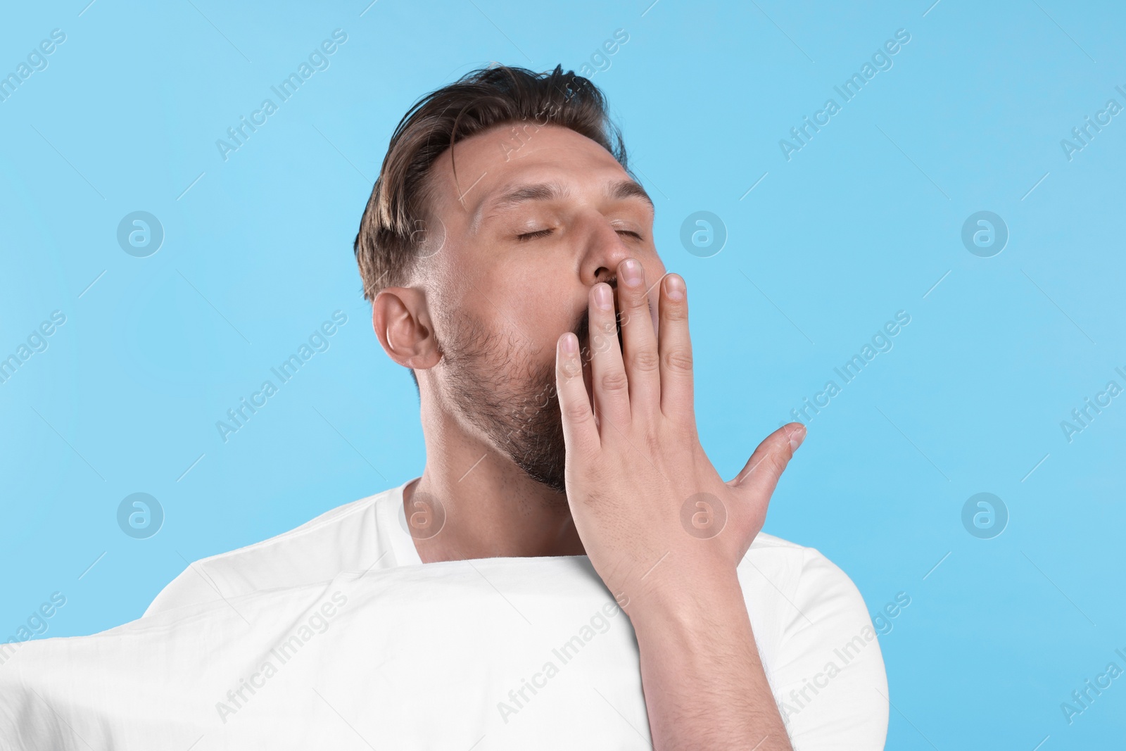 Photo of Sleepy man with pillow yawning on light blue background. Insomnia problem