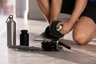 Photo of Male athlete assembling adjustable dumbbell near bottle, parts and jar of protein on floor indoors