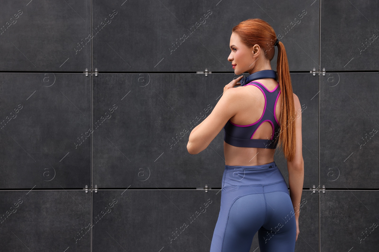 Photo of Beautiful woman in stylish gym clothes with headphones posing near dark grey wall on street, space for text