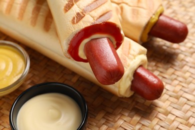 Photo of Tasty french hot dogs and dip sauces on wicker mat, closeup