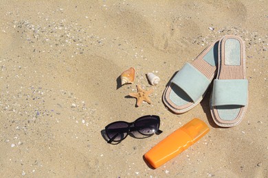 Photo of Stylish sunglasses, slippers and bottle of sunblock on sand, space for text. Beach accessories