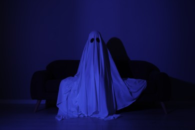 Photo of Creepy ghost. Woman covered with sheet sitting on sofa in blue light