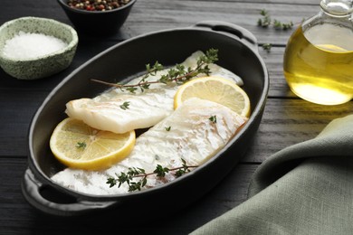 Photo of Fresh raw cod fillets with thyme and lemon in baking dish on black wooden table