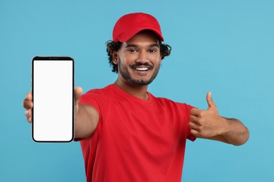 Image of Happy courier holding smartphone with empty screen and showing thumbs up on light blue background