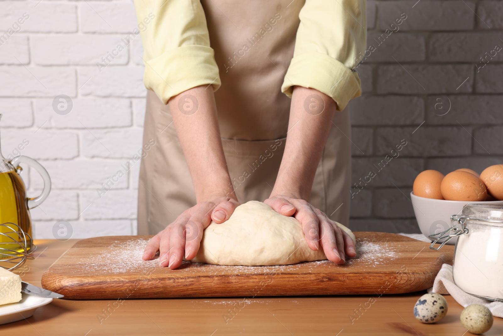 Photo of Woman kneading dough at wooden table near white brick wall, closeup