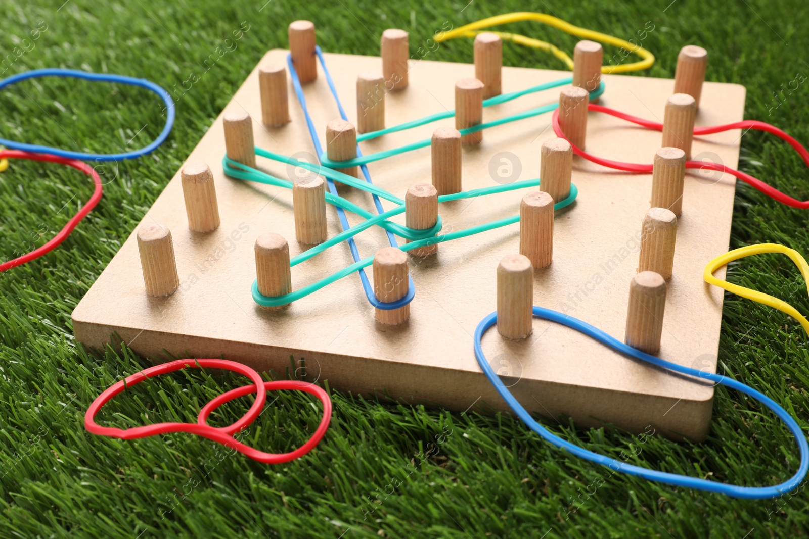 Photo of Wooden geoboard with rubber bands on artificial grass, closeup. Educational toy for motor skills development