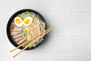Photo of Bowl of delicious rice noodle soup with meat and egg on white tiled table, top view. Space for text