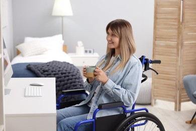 Woman in wheelchair with cup of drink using computer at home