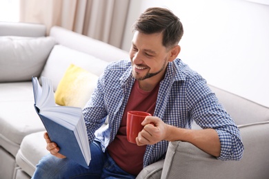 Photo of Handsome man with cup of coffee reading book on sofa at home