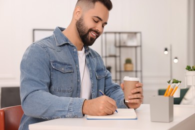 Young man with cup of coffee writing in notebook at white table indoors